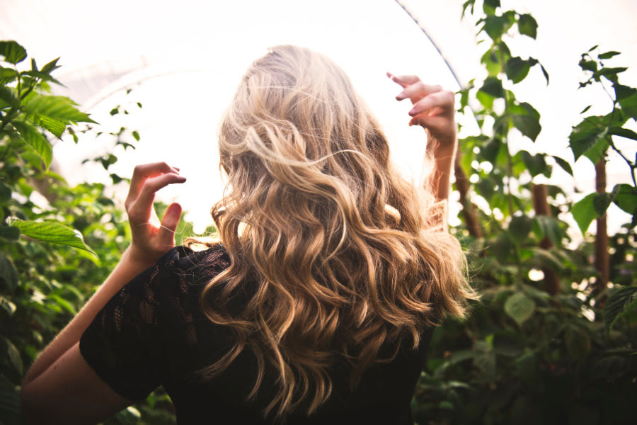 Kokosöl: Ein unterschätztes Multitalent 4 Blonde Frau mit langen welligen Haaren steht von hinten inmitten grüner Pflanzen im warmen Sonnenlicht.