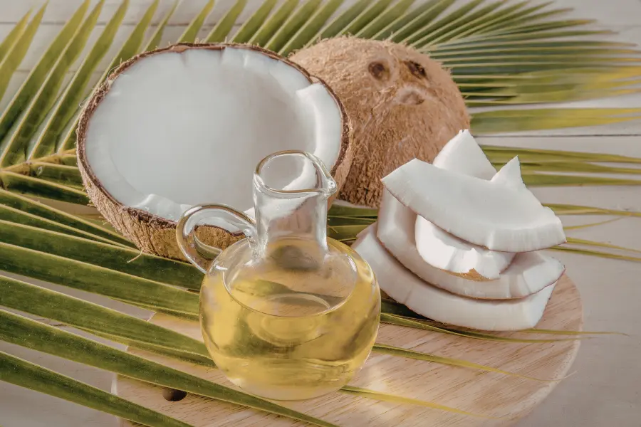 Kokosöl: Ein unterschätztes Multitalent 2 Kokosnussöl in einer Glasflasche mit halbierter Kokosnuss, frischen Kokosstücken und grünen Palmblättern auf einem hellen Holzbrett.