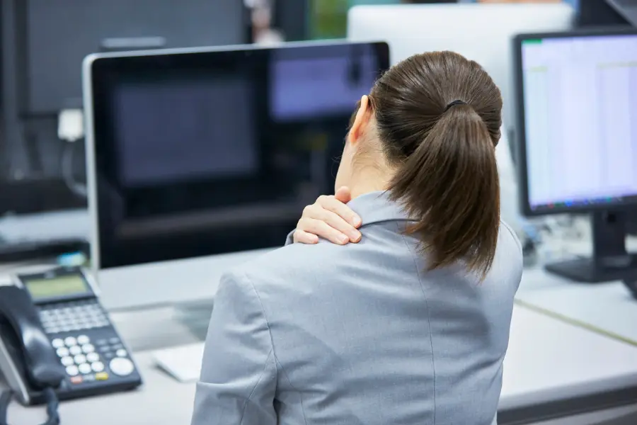 Frau im Büro massiert ihren Nacken, während sie vor mehreren Computerbildschirmen sitzt – Symbolbild für Nackenschmerzen und Verspannungen am Arbeitsplatz.