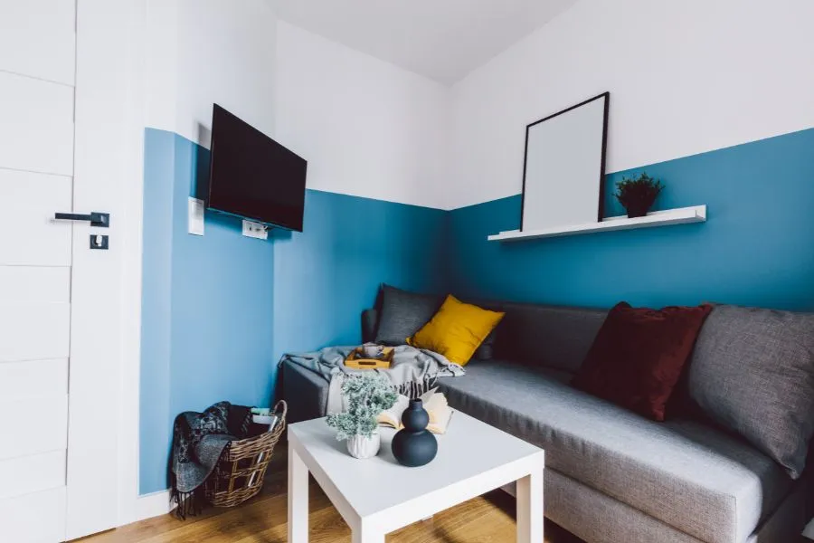 Rabatte Coupons Kleines Wohnzimmer mit blauer Wand, grauem Sofa, kleinem Couchtisch, bunten Kissen und einem schmalen Wandregal unter einem Fernseher.