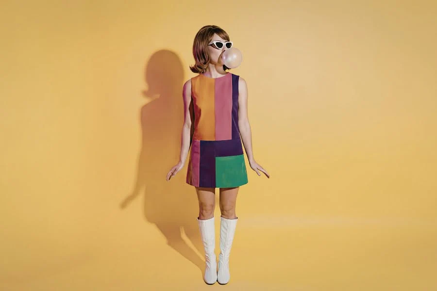 Ihr seht eine Frau in einem bunten, geometrischen Kleid mit Sonnenbrille, die vor einem gelben Hintergrund Kaugummi bläst.