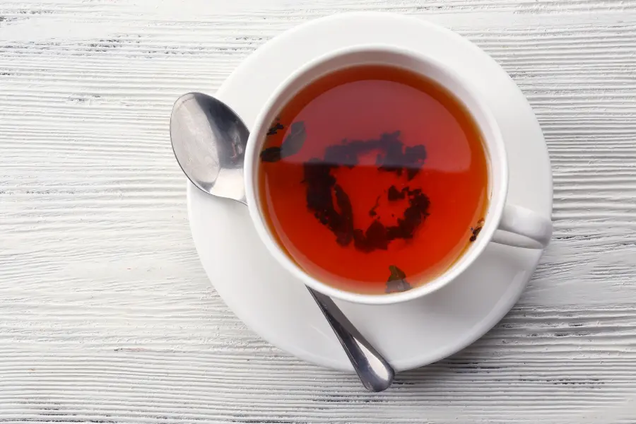 Tasse mit frisch aufgebrühtem schwarzem Tee und sichtbaren losen Teeblättern auf weißem Holztisch.
