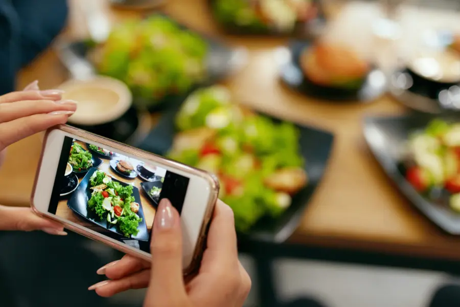 Mehr Leute wollen dank social Media gesunde Ernährung, unterstützt kann das mit einem Sundaynatural Gutschein werden. Person fotografiert mit einem Smartphone mehrere Salate und Speisen auf einem gedeckten Tisch.