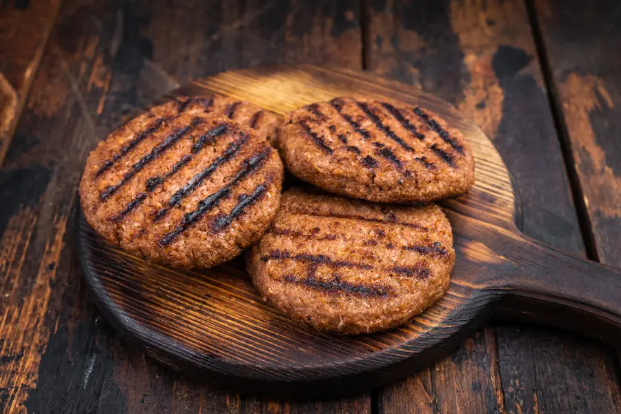 Büffel Bill Gutschein sichern und sparen: so gut kann gesunde Ernährung sein. Drei gegrillte Burger-Patties mit Grillstreifen liegen auf einem rustikalen Holzbrett.