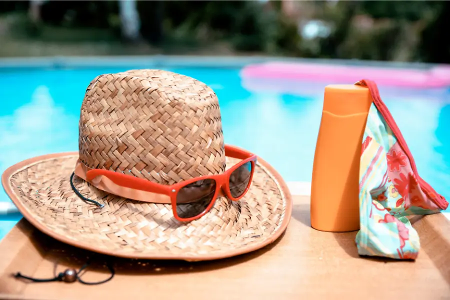 Bei Shop-Apotheke findet ihr immer hochwertige Produkte, die eure Haut schutzt und dafür sorgt braun werden ohne Risiko zu können. Strohhut mit Sonnenbrille, Sonnencreme und bunter Badebekleidung liegen auf einem Tisch am Pool.