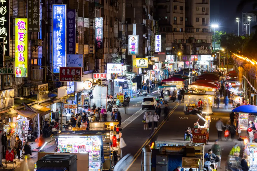 Busy Asian night market street with illuminated shop signs, food stalls, traffic, and crowds of people.
