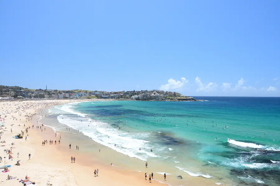 Wide view of a crowded sandy beach with turquoise ocean waves, swimmers, and a clear blue sky.