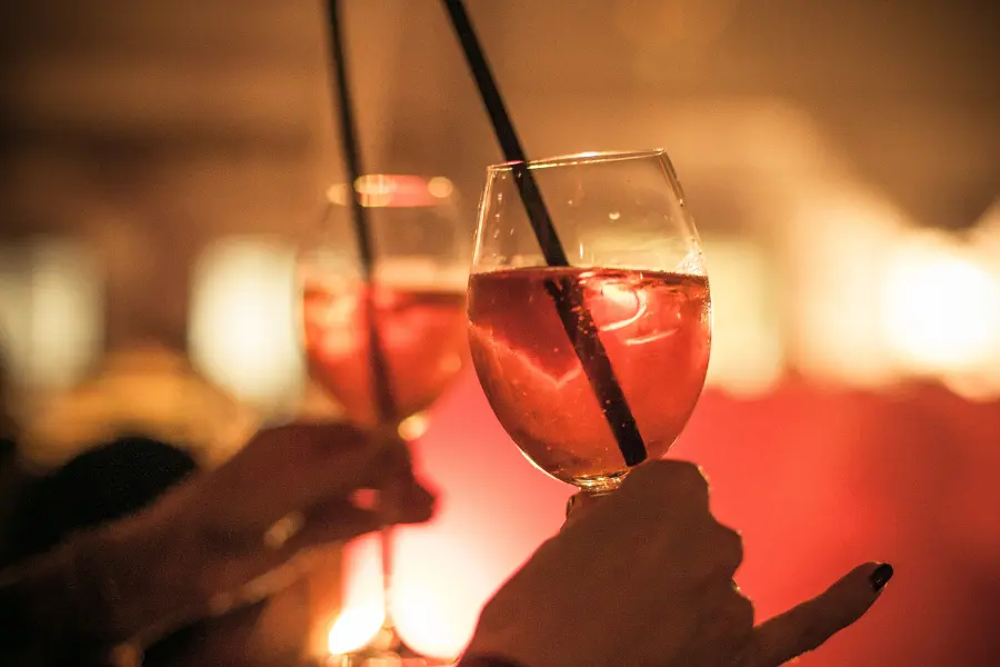 Two people holding glasses of pink cocktails with straws in a warm, softly lit bar or lounge setting.