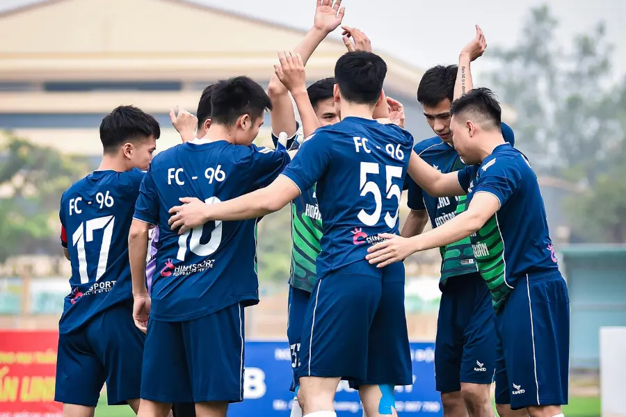 Soccer players in blue uniforms huddle together and raise their hands before a match on an outdoor field.