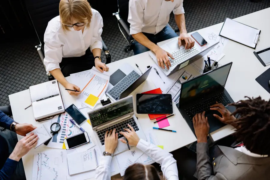 Team of professionals working together around a shared office table with laptops, documents, charts, notebooks, and digital devices.