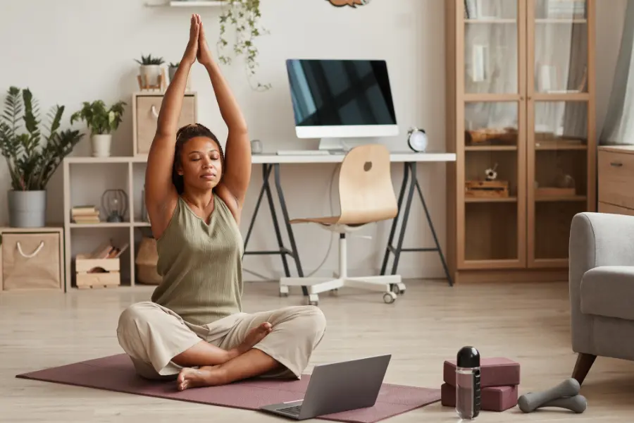 Woman practicing yoga on a mat at home with a laptop, water bottle, yoga blocks, and dumbbells nearby.