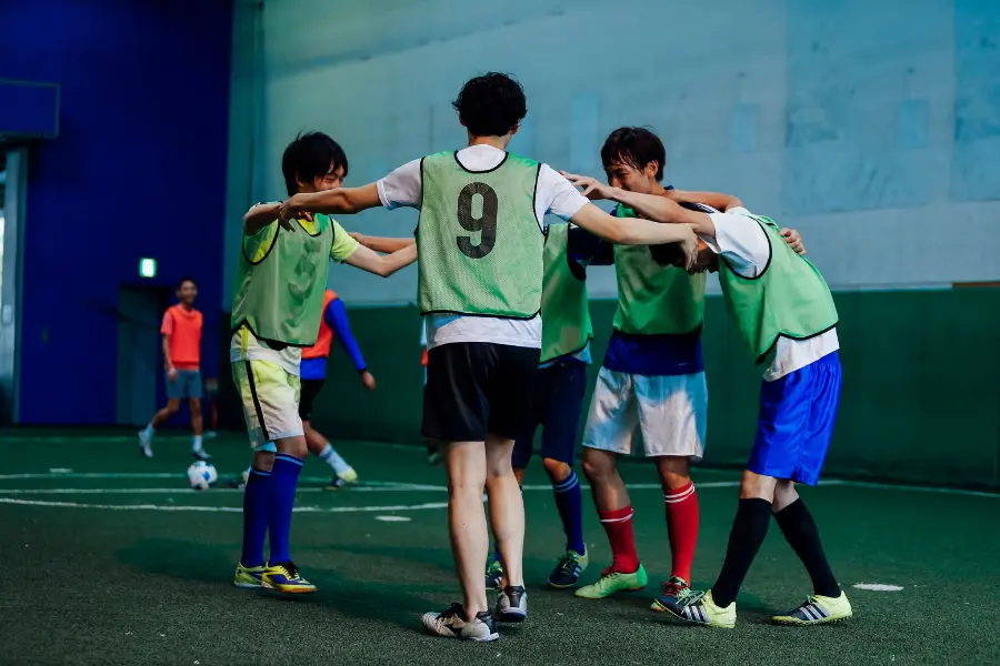 Indoor soccer players wearing green training bibs huddle together with arms around each other during a team practice session.