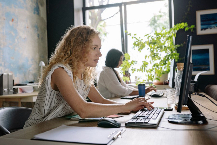 Two women working at desktop computers in a modern office with natural light, plants, and shared workspace desks.
