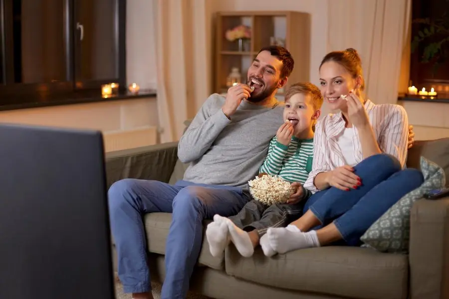 Eine dreiköpfige Familie sitzt gemütlich auf einem Sofa, schaut gemeinsam fern und isst Popcorn. Der Raum ist stimmungsvoll mit Kerzen beleuchtet, die Atmosphäre wirkt entspannt und wohnlich.