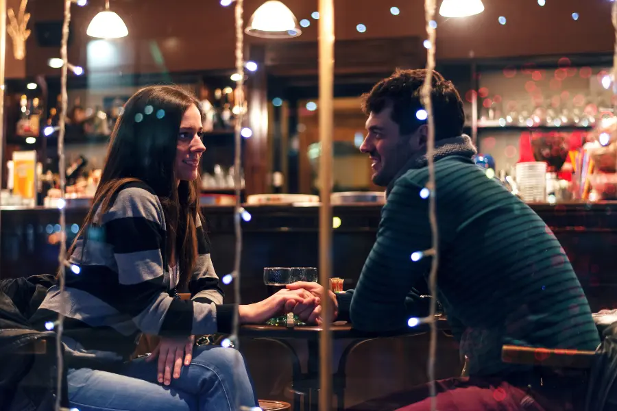Der LaBlue Gutschein unterstützt die Partnersuche im Internet. Couple smiling and holding hands during a romantic date in a warmly lit restaurant with decorative string lights.