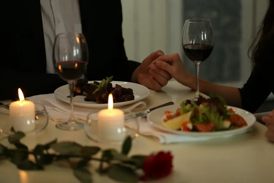 Der Lovepoint Gutschein gewährt euch Einlass in die Partnersuche im Internet. Couple holding hands during a romantic candlelit dinner with wine glasses, plated food, and a red rose on the table.