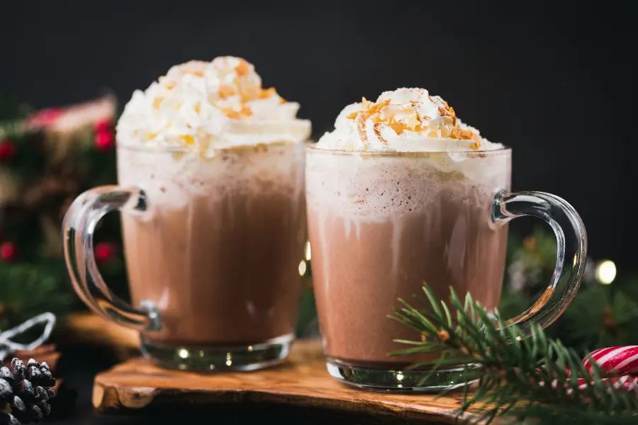 ☕️ 3 leckere Wintergetränke, die euer Herz erwärmen 2 Two mugs of hot chocolate topped with whipped cream and toasted coconut, surrounded by festive greenery and holiday decorations.