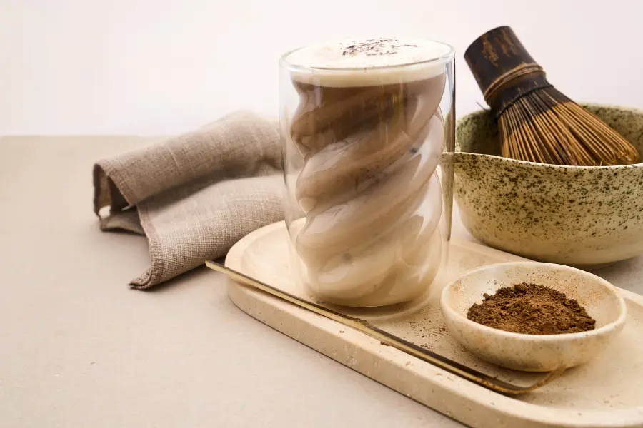 ☕️ 3 leckere Wintergetränke, die euer Herz erwärmen 1 Creamy mat cha latte in a double-walled glass served with mat cha powder, a bamboo whisk, and a ceramic bowl on a neutral tabletop.