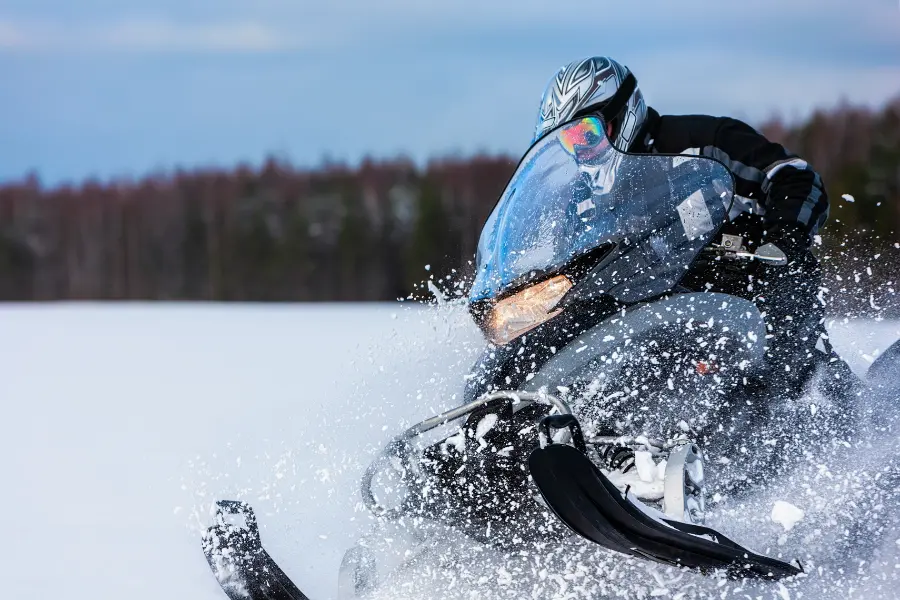 Mit dem The North face Gutschein erhalten jetzt alle Studenten 10% Rabatt auf die Bestellung. Person fährt mit einem Schneemobil durch tiefen Schnee und wirbelt dabei Schnee vor einer winterlichen Waldkulisse auf.