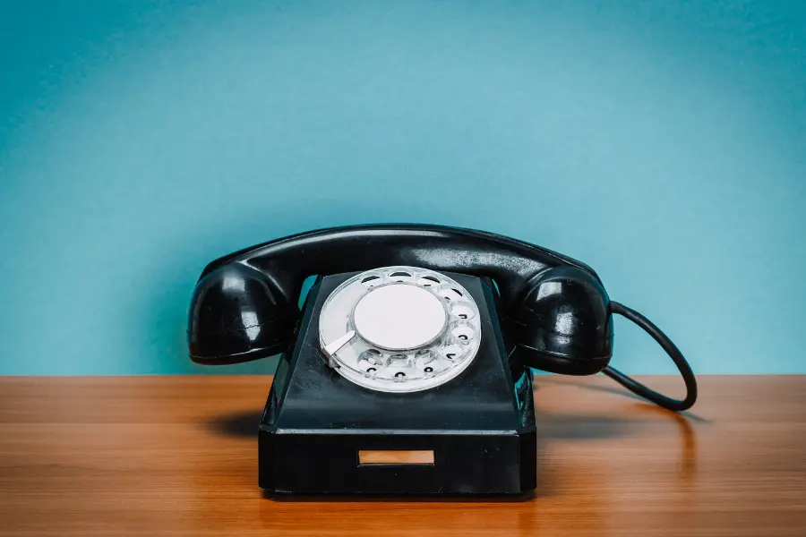 Vom Wählscheibentelefon bis zum modernen Smartphone - die Entwicklung ging immer richtung portabler und leichter bedienbar. Vintage black rotary telephone on a wooden table against a blue background.