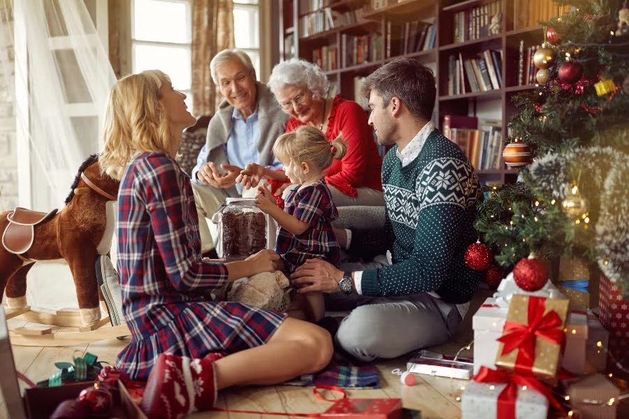 Eine Familie sitzt vor einem geschmückten Weihnachtsbaum im Wohnzimmer. Zwei ältere Personen, zwei Erwachsene und ein Kind befinden sich im Kreis auf dem Boden. Das Kind hält ein Geschenk in den Händen, während die anderen zuschauen. Um sie herum liegen weitere verpackte Geschenke, und im Hintergrund stehen Bücherregale sowie ein Schaukelpferd.