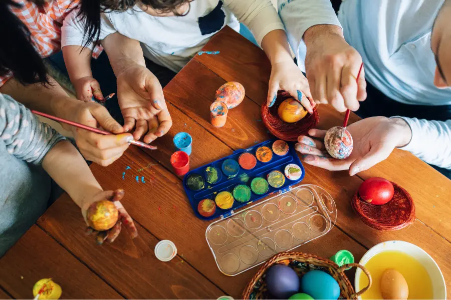 Mehrere Personen bemalen gemeinsam Ostereier an einem Holztisch mit Pinseln und bunten Farben.