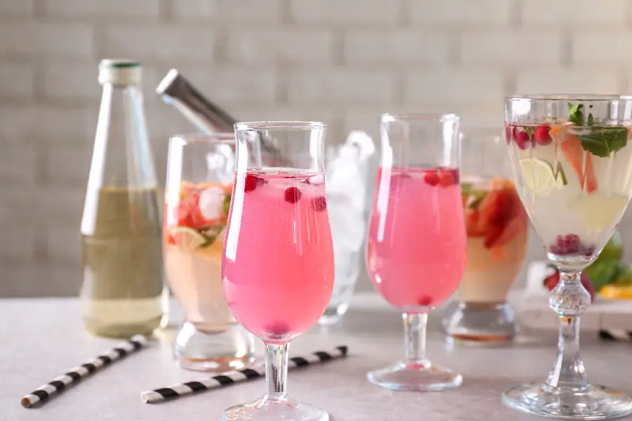 Mehrere Gläser mit bunten, alkoholfreien Cocktails in Rosa- und Gelbtönen, garniert mit Beeren, Minze und Eiswürfeln – daneben eine Flasche mit heller Flüssigkeit auf hellem Untergrund.