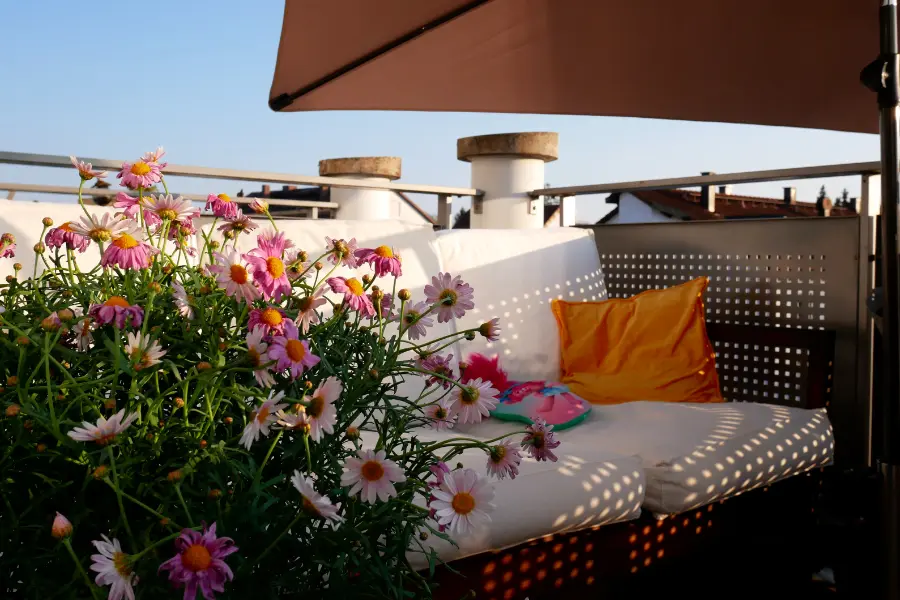 Gemütliche Dachterrasse mit rosa Gänseblümchen, heller Sitzbank, gelbem Kissen und Sonnenschirm im warmen Abendlicht.