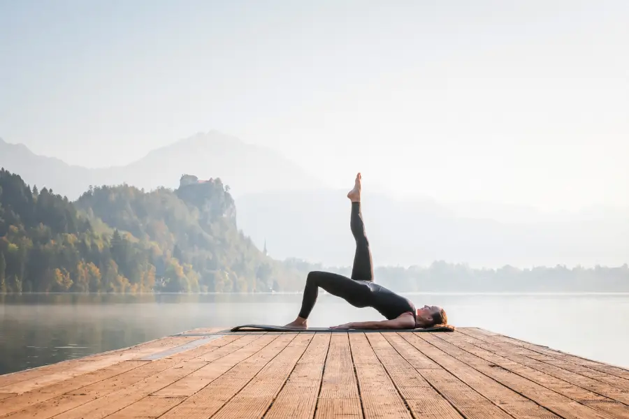 Im Einklang mit der Natur: Eine Frau praktiziert eine Yogaübung am stillen Seeufer. Die Übung symbolisiert Ausgeglichenheit, innere Ruhe und die Verbindung von Körper und Geist – fern vom Alltagstrubel. Eine Frau macht Yoga auf einem Holzsteg an einem ruhigen See, umgeben von Bergen und Morgennebel. Sie liegt auf der Matte und streckt ein Bein senkrecht nach oben – eine entspannte und kraftvolle Pose.