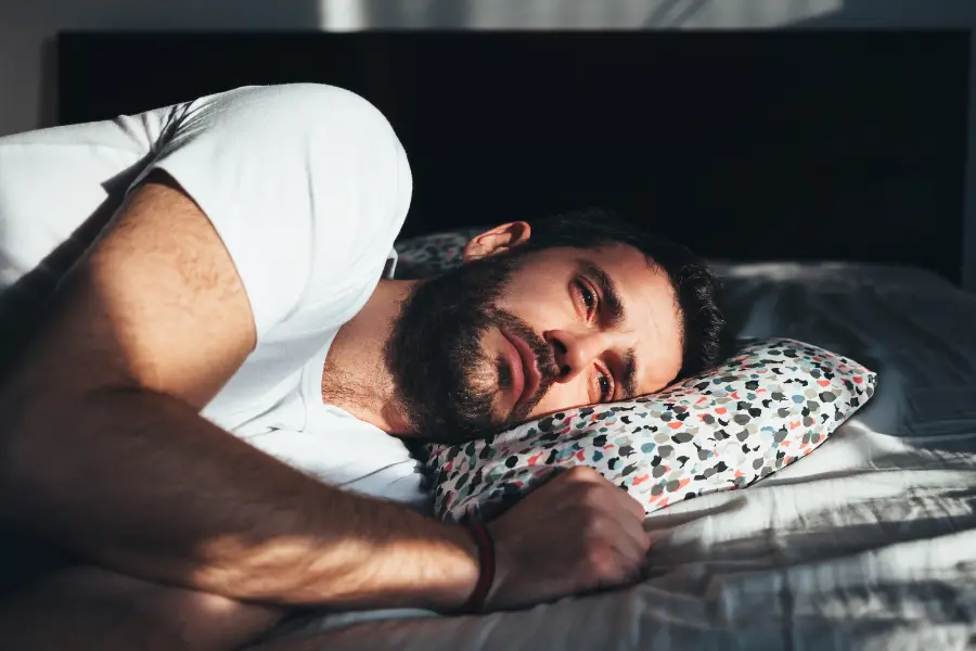 Das Bild zeigt einen jungen Mann, der mit offenen Augen und ruhigem Blick auf der Seite liegt. Die Szene vermittelt einen Zustand innerer Unruhe oder gedanklicher Überforderung – ein Moment der Reflexion oder möglicher Schlaflosigkeit. Ein nachdenklich wirkender Mann liegt seitlich auf einem Bett, den Kopf auf ein bunt gemustertes Kissen gestützt, während Sonnenlicht durch das Fenster fällt.