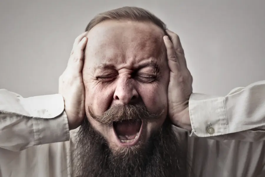 Ein bärtiger Mann mit geschlossen Augen und schmerzverzerrtem Gesicht schreit laut, während er sich mit beiden Händen die Ohren zuhält.