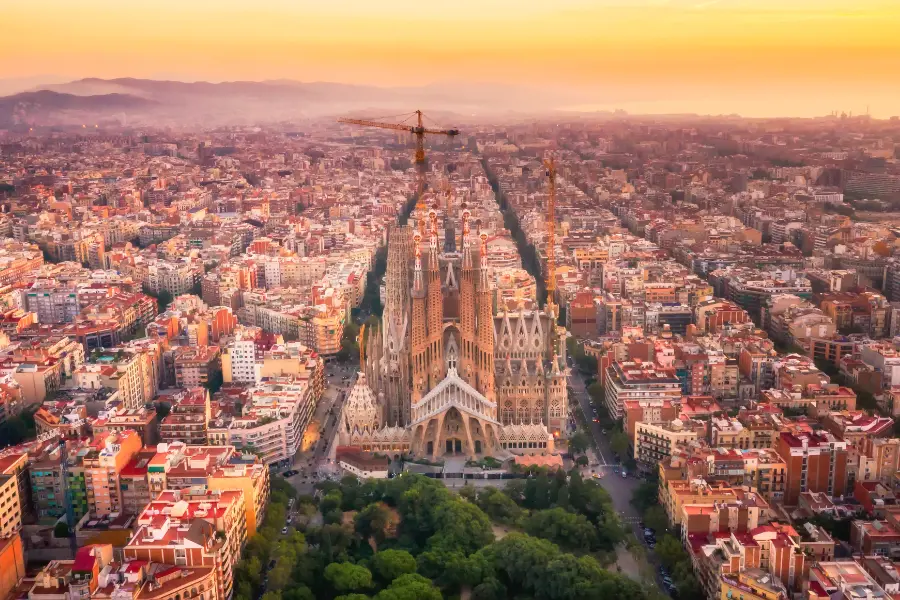 Luftaufnahme der Sagrada Família im Herzen Barcelonas mit Baukränen zwischen dicht bebauten Stadtvierteln der spanischen Metropole im goldenen Licht.