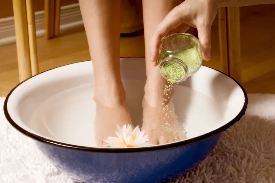 Füße pflegen - diese Tipps garantieren strahlende Sommerfüße | www.rabatt-coupon.com Zwei Füße in einer Schüssel mit warmem Wasser, während grüne Badesalze hineingestreut werden – daneben eine dekorative Blüte.