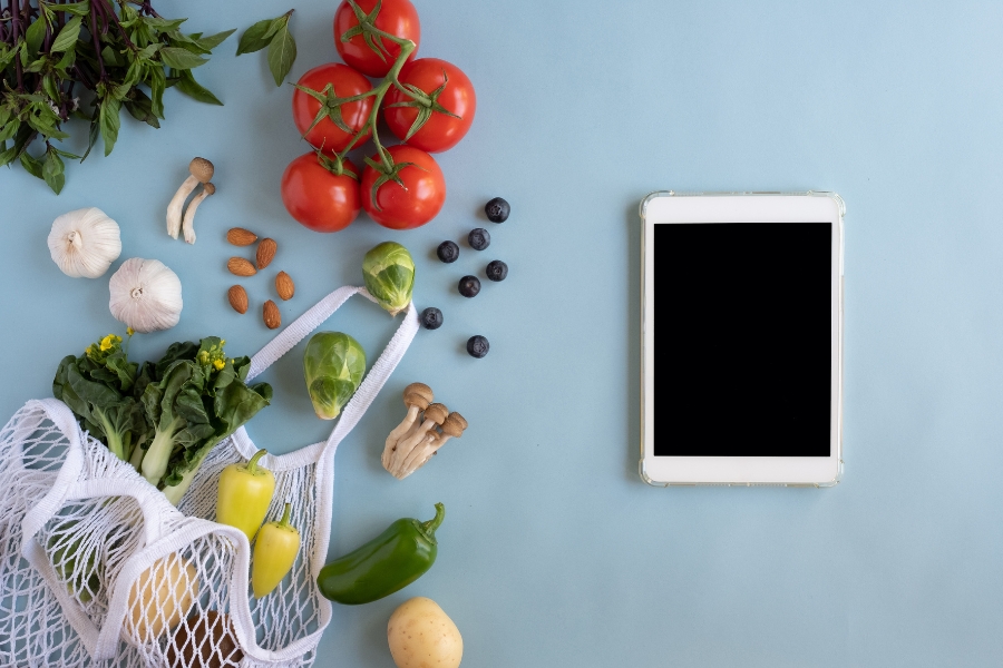 Nachhaltig Einkaufen: Onlineshopping im Einklang mit dem veganen Lifestyle 🌱 12 Tomaten, Früchte, Gemüse, Pilze, Tasche und Tablet auf blauen Hintergrund