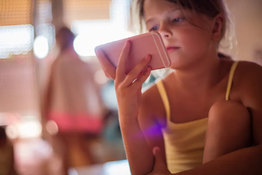 Ein Mädchen im gelben Top sitzt in einem abgedunkelten Raum und schaut konzentriert auf ein rosafarbenes Smartphone – die Umgebung ist unscharf, im Hintergrund erkennt man andere Kinder.