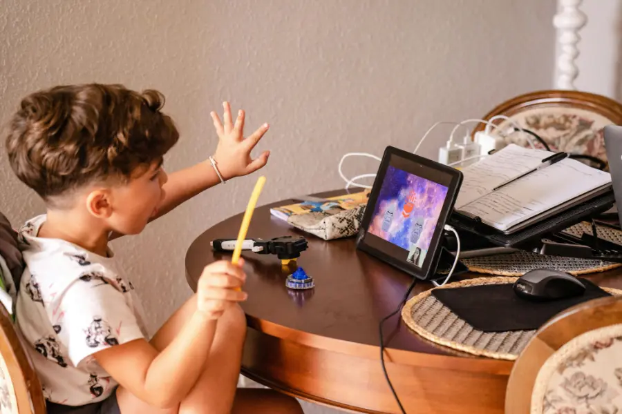 Nutzt den O2 Gutschein, um euren Kindern Sicherheit und Kontrolle im Internet zu geben. Kind sitzt an einem Tisch mit einem Tablet, hebt die Hand und hält einen Bleistift – im Hintergrund Schulmaterialien, Hefte und eine Computermaus. Es scheint an einem interaktiven Unterricht oder Spiel teilzunehmen.