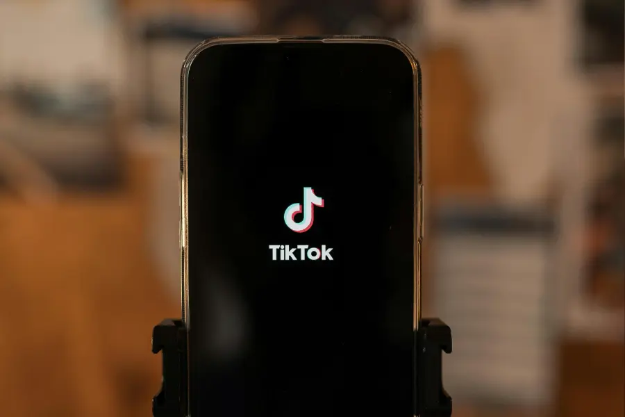 Das Internet bietet den Kindern nicht nur Spiel und Spaß. Es lauern auch Gefahren, vor denen wir sie beschützen müssen. Smartphone mit TikTok-Startbildschirm im Vordergrund, auf einem Stativ montiert. Der Hintergrund ist unscharf und warm beleuchtet.
