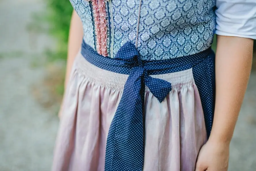 O'zapft is! - das richtige Outfit zum Oktoberfest 2025 5 Zu sehen ist der Mittelteil eines klassischen Dirndls mit rosafarbener Schürze und gemustertem Mieder in Blau. Die Schleife ist links gebunden – traditionell ein Hinweis darauf, dass die Trägerin ledig ist. Das Bild zeigt keine Person im Ganzen, sondern fokussiert auf das Detail der gebundenen Schürze.