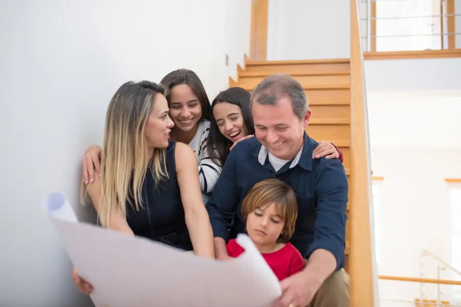Hausbauen so teuer wie noch nie - So spart ihr richtig 2 Familie mit Bauplan auf Treppe sitzend, lächelnd und gemeinsam die Planung ihres zukünftigen Hauses betrachtend