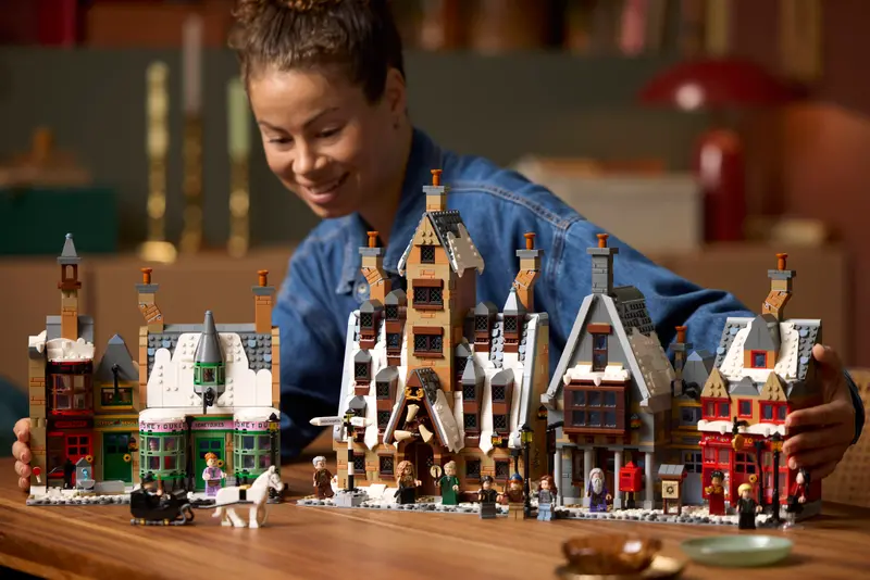 LEGO begeistert im Herbst – Von Hogwarts bis zur IAA – Die spannendsten Neuheiten im Überblick 10 Frau spielt mit dem LEGO Harry Potter Hogsmeade Set auf einem Holztisch. Die verschneiten Häuser und Minifiguren wie Harry, Hermine und Ron sind detailreich aufgebaut und versetzen in die magische Welt von Hogwarts. Die Szene ist warm beleuchtet und zeigt Begeisterung für kreativen Bauspaß.