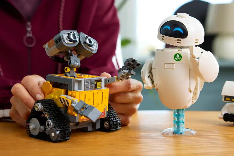 LEGO begeistert im Herbst – Von Hogwarts bis zur IAA – Die spannendsten Neuheiten im Überblick 9 Das Bild zeigt zwei liebevoll gestaltete LEGO-Modelle der berühmten Pixar-Charaktere WALL·E und EVE aus dem Animationsfilm WALL·E. Die detailreichen Figuren stehen auf einem Holztisch und werden von einer Person mit dunkelroter Jacke in Szene gesetzt.