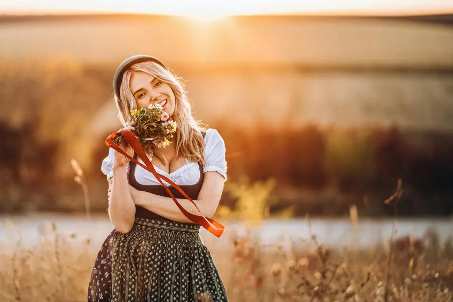 O'zapft is! - das richtige Outfit zum Oktoberfest 2025 7 Frau im Dirndl mit Blumenstrauß auf einem Feld bei Sonnenuntergang – bayerische Tradition und Lebensfreude im Spätsommer.