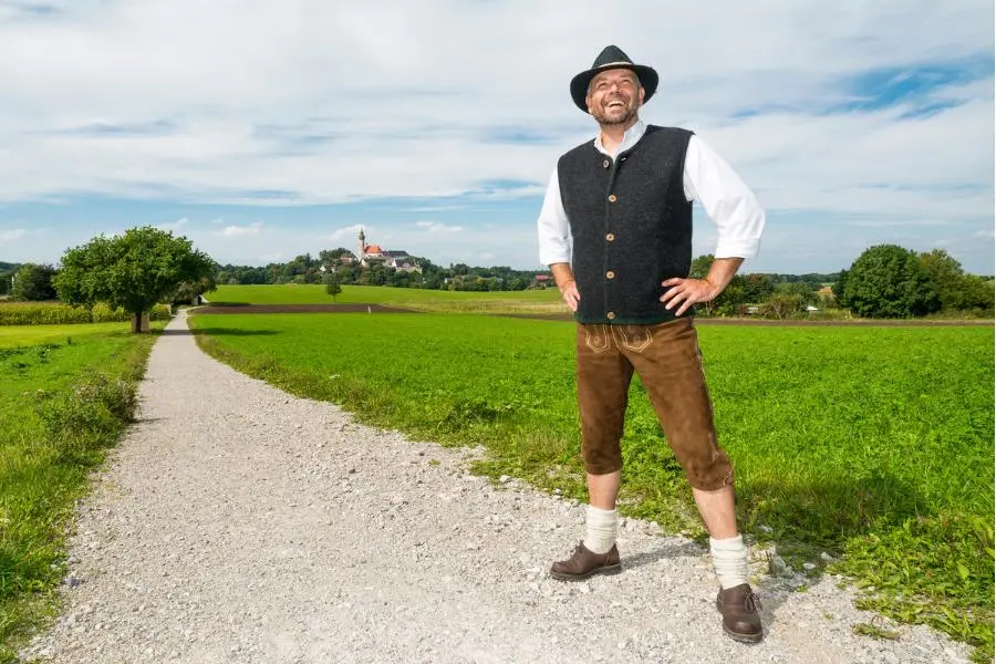 O'zapft is! - das richtige Outfit zum Oktoberfest 2025 6 Mann in bayerischer Tracht mit Lederhose und Weste steht auf Feldweg vor idyllischer Landschaft mit Kirche im Hintergrund.