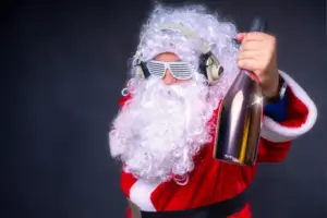 Ein verrückter Weihnachtsmann mit weißem Rauschebart, stylischer Partybrille und Kopfhörern hält eine Champagnerflasche in die Kamera – bereit für die Weihnachtsfeier.