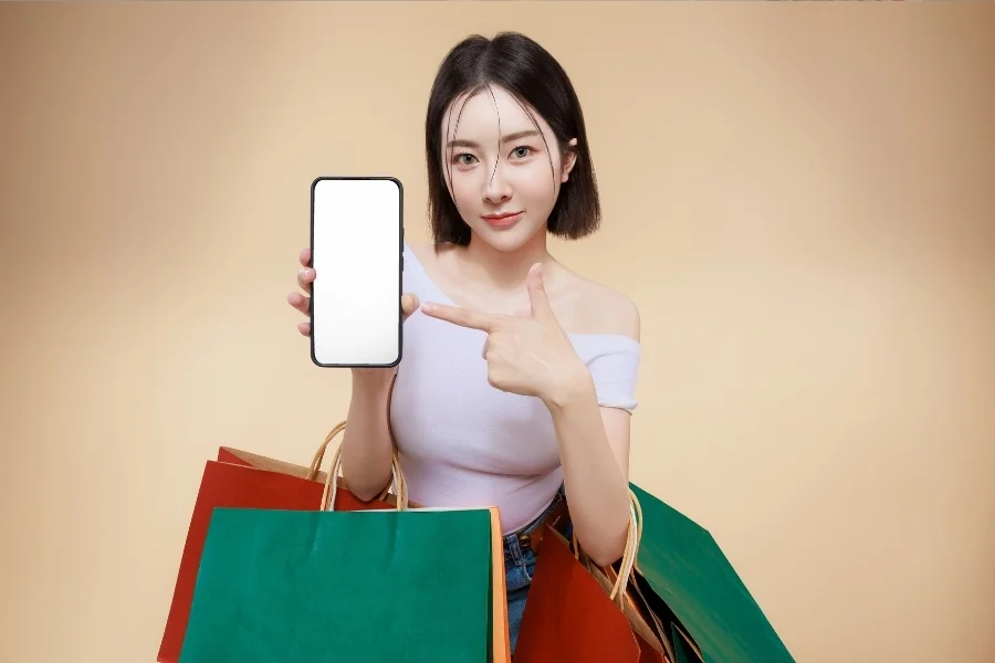 Frau hält Smartphone mit leerem Bildschirm und zeigt darauf während sie mehrere Einkaufstaschen trägt